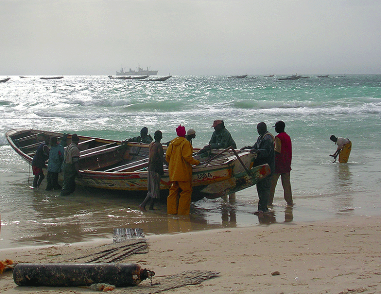 Retour de mer pour cette grande pirogue qui va bientôt être remontée en haut de la plage, probablement à l’aide de bouteilles de gaz – comme celle du premier plan – en guise de rouleaux. Remarquer les nombreux bateaux restés au mouillage au-delà des brisants.