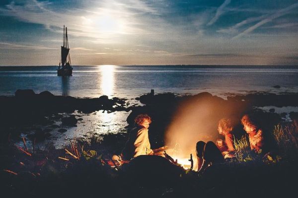 Plusieurs membres de l'équipage, faisant un feu à terre, avec le bateau en mer, ancrée.